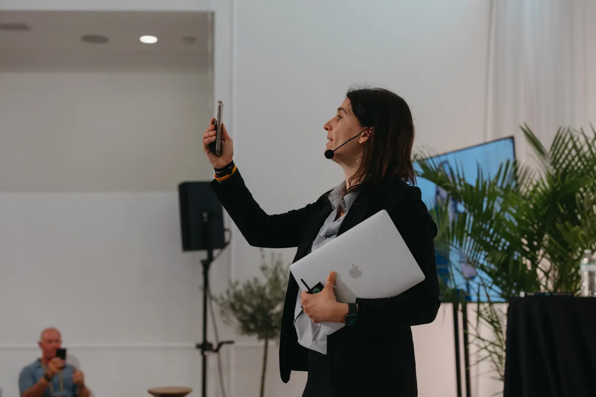 Dr. Ariana on stage holding a phone in one hand and a MacBook in the other — demonstrating the device-load every knowledge worker carries.